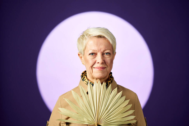volwassen blonde vrouw met beige ventilator door haar borst staan tegen grote volle maan over lavendel achtergrond en kijken naar camera - Foto, afbeelding
