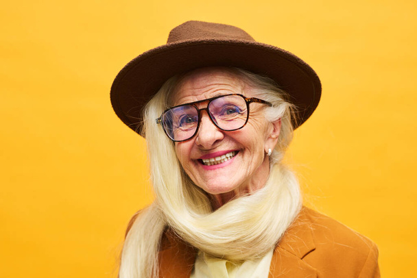 Nahaufnahme Porträt einer fröhlichen Seniorin mit braunem Fedora-Hut und Brille, deren lange weiße Haare um den Hals gebunden sind - Foto, Bild