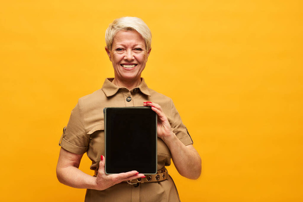 Vrolijke volwassen vrouw met kort blond haar holding tablet met leeg scherm, terwijl het maken van de presentatie van project punten in isolatie - Foto, afbeelding