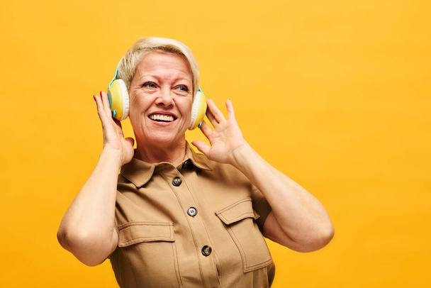 Fröhliche reife Frau mit kurzen blonden Haaren genießt Musik mit Kopfhörern, während sie isoliert vor der Kamera steht - Foto, Bild