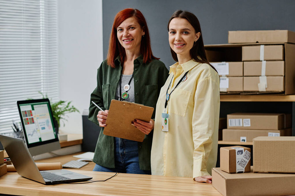 Two successful female volunteers standing by workplace in storage room and looking at camera while working with online information - Foto, Imagen