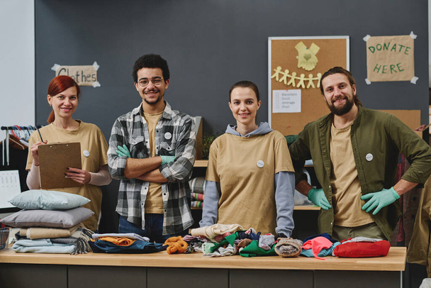 Group of happy young intercultural volunteers looking at camera with smiles while standing by table with stacks of second hand clothes - Фото, зображення