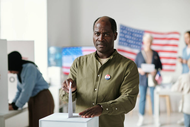 Mature black man looking at camera while putting paper with his choice of candidate into ballot box while taking part in presidential elections - Photo, Image