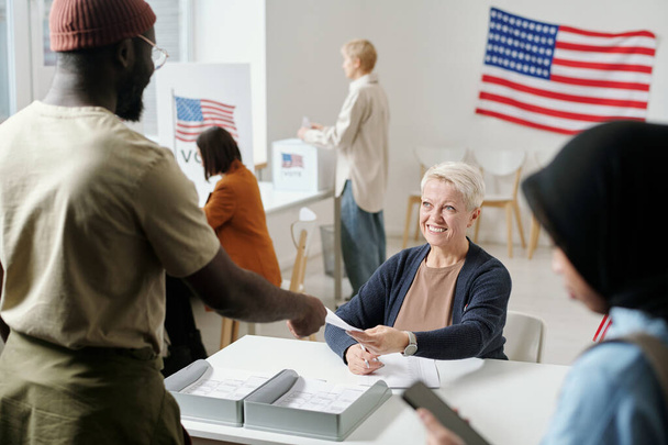 Щаслива білява зріла жінка передає бюлетень молодому афроамериканцю, що стоїть за виборчою комісією. - Фото, зображення