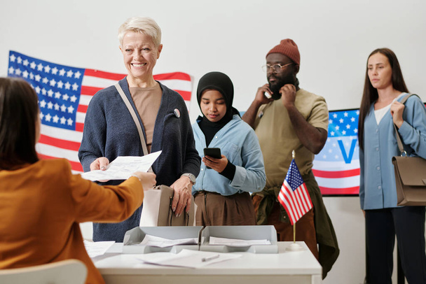 Очередь межкультурных избирателей, стоящих у стола избирательной комиссии, в то время как один из них берет избирательный бюллетень со списком кандидатов - Фото, изображение