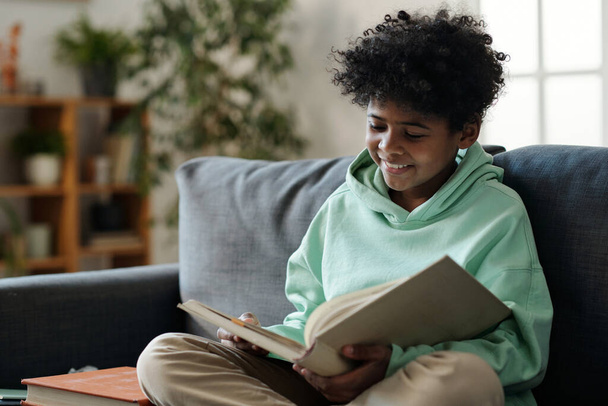 Écolier afro-américain souriant en tenue décontractée lisant le livre assis sur le canapé dans le salon et se préparant à la leçon de littérature - Photo, image