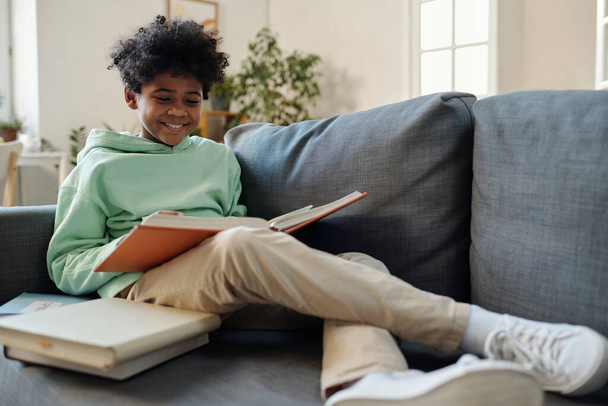 Adolescent écolier en causalvêtements assis sur le canapé et la lecture de l'encyclopédie tout en se préparant pour la leçon de géographie ou de biologie - Photo, image