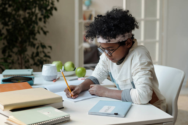 Inspired African American schoolboy writing essay or drawing in copybook while sitting by desk in living room and preparing homework - Fotó, kép