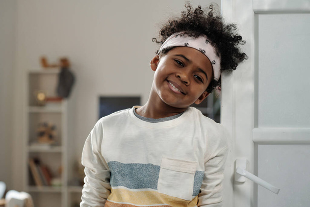 Boldog imádnivaló iskolás gyerek fejpántban és alkalmi pulóverben, mosolyogva néz a kamerába, miközben a nappali belső terével szemben áll. - Fotó, kép