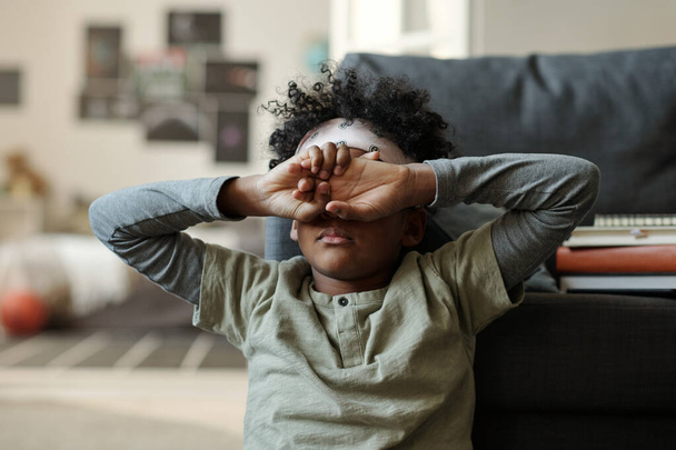 Offended or tired African American boy in grey pajamas hiding his face with hands while sitting by couch on the floor of living room - Фото, зображення