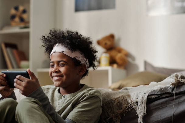 Sorrindo menino afro-americano na cabeça olhando para a tela do smartphone enquanto se comunica em vídeo chat ou assistindo filme online - Foto, Imagem
