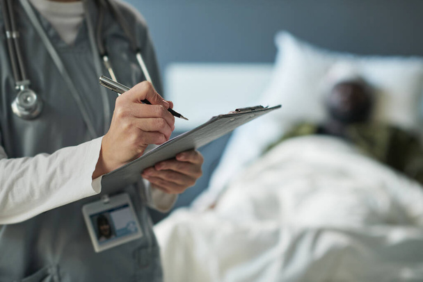 Mão de jovem enfermeira com caneta sobre o documento médico fazendo prescrições enquanto está de pé contra o soldado ferido na cama - Foto, Imagem