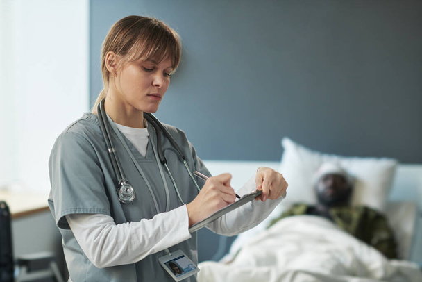 Verpleegster in uniform maakt aantekeningen in medisch document terwijl ze voor de camera staat tegen gewonde soldaat in militair ziekenhuis - Foto, afbeelding