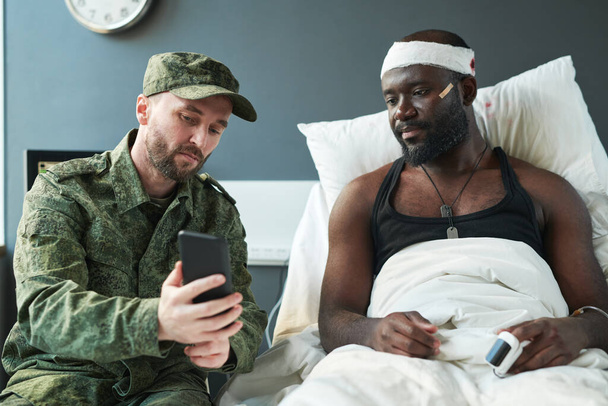 Young soldier in military uniform showing new photos in smartphone to injured friend with bandaged head sitting on bed in hospital ward - Foto, afbeelding