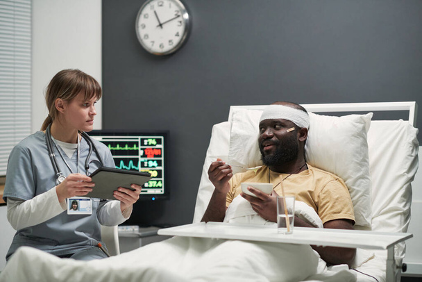 Blessé Afro-Américain avec tête pansée mangeant le petit déjeuner au lit et parlant à l'infirmière entrant les données dans la tablette dans la salle d'hôpital - Photo, image