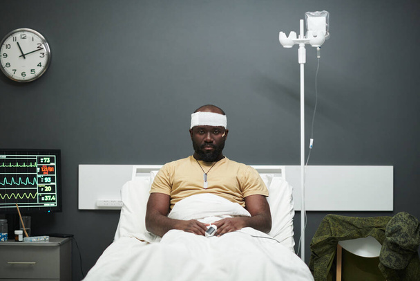 Молодий афроамериканець з оксиметром на кінчику пальця і пов'язкою на голові сидячи в ліжку в лікарняній палаті і дивлячись на камеру - Фото, зображення