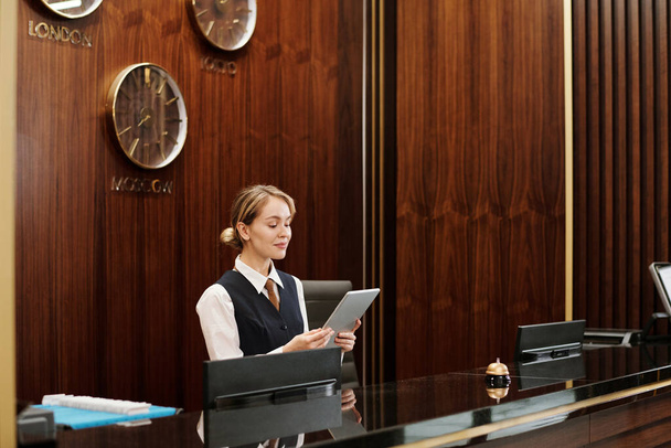 Νεαρή ξανθιά γυναίκα ρεσεψιονίστ σε ομοιόμορφη κοιτάζοντας οθόνη tablet, ενώ στέκεται δίπλα μετρητή στο σαλόνι του σύγχρονου ξενοδοχείου και τη δικτύωση - Φωτογραφία, εικόνα