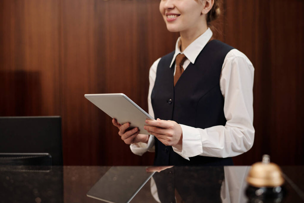 Close-up van een jonge vrouw in uniform van een receptioniste die een tablet vasthoudt terwijl zij voor de camera op de werkplek staat en op de gasten wacht - Foto, afbeelding