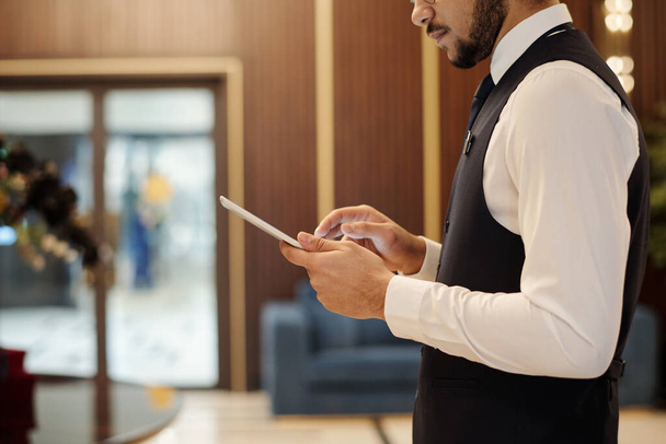 Lüks otellerde çalışan, dijital tablet kullanan genç bir erkek çalışanın yan görüntüsü geniş bir salonda kameranın önünde duruyor. - Fotoğraf, Görsel