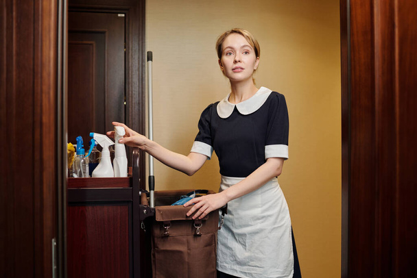 Young pretty room maid in uniform taking bottle with sprayer from cart while standing by open door in corridor of luxurious hotel - Foto, afbeelding