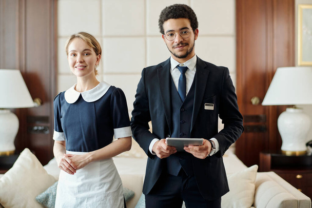 Young successful staff of luxurious five star hotel standing against couch with cushions and two lamps in room prepared for new guests - Foto, imagen
