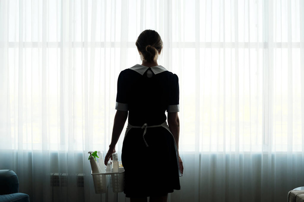 Widok z tyłu pokojówka z detergentami w koszu idąc w kierunku dużego okna pokryte białymi zasłonami szyfonu w pokoju hotelowym - Zdjęcie, obraz