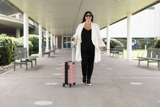 Reife Frau in weißem Mantel verlässt Terminal und trägt ihren pinkfarbenen Koffer auf Rädern. Konzept der Geschäftsreise. Konzept der Geschäftsreise. - Foto, Bild