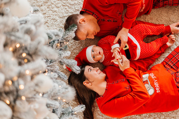 Christmas. Family. Happiness. Top view of dad, mom and daughter in Santa hats looking at camera and smiling while lying on the floor at home. - Фото, изображение