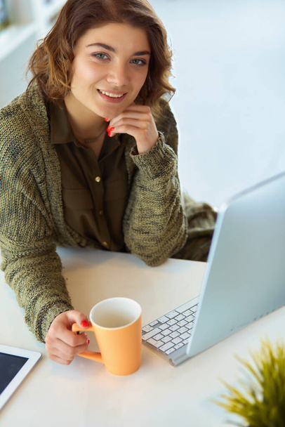 Attractive smiling woman sitting at office desk, holding a cup of coffee, she is relaxing and looking away. - Foto, Bild