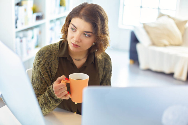 Attractive smiling woman sitting at office desk, holding a cup of coffee, she is relaxing and looking away. - Foto, imagen