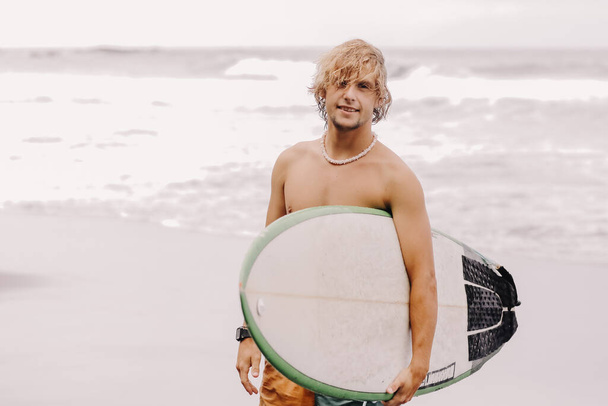 Handsome fit young blond man with mock up surfboard waits for wave to surf spot at sea ocean beach with black sand and looks at camera. Concept of sport, fitness, freedom, happiness, new modern life. - Фото, изображение