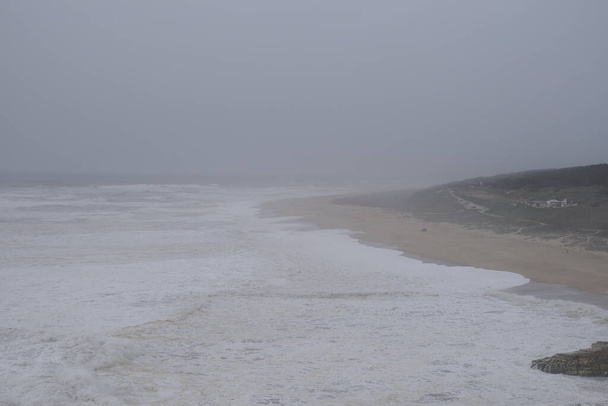Poziome zdjęcie ponurego burzliwego morza, szorstkie fale, peleryna pokryta lasem widoczna jest w oddali. Piankowe fale toczą się wzdłuż piaszczystego brzegu. Wieczór. Niebo Zmierzchu, Portugalia, Nazar - Zdjęcie, obraz
