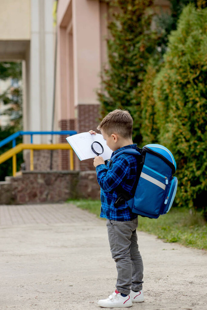 A little schoolboy looking through a magnifying glass at a textbook. Education for young children. Back to school concept. - Zdjęcie, obraz