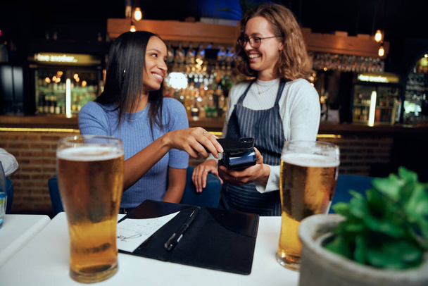 Happy young woman wearing casual clothing paying for drinks with wireless technology next to waitress - Foto, Imagem