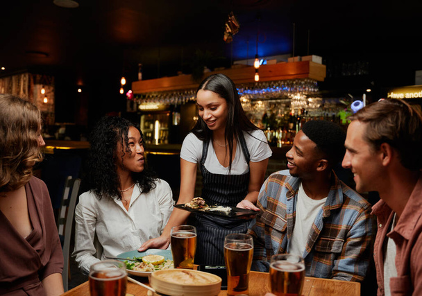 Glückliche junge multiethnische Gruppe von Freunden in lässiger Kleidung, die das Abendessen von der Kellnerin an der Bar erhalten - Foto, Bild