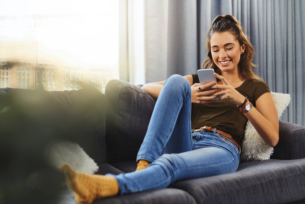 Finally getting a chance to reply to some messages. Full length shot of a beautiful young woman using a cellphone while relaxing on a couch at home - Fotografie, Obrázek