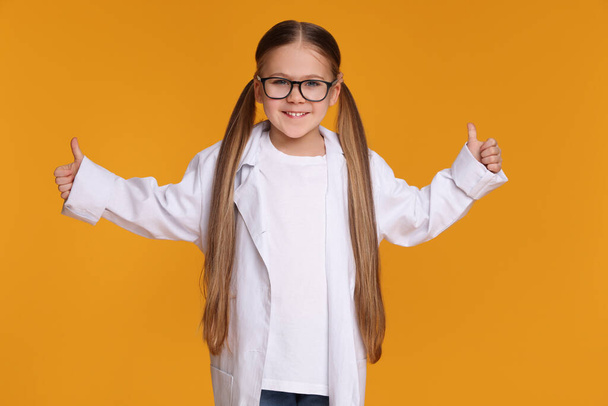 Little girl in medical uniform showing thumbs up on yellow background - Zdjęcie, obraz