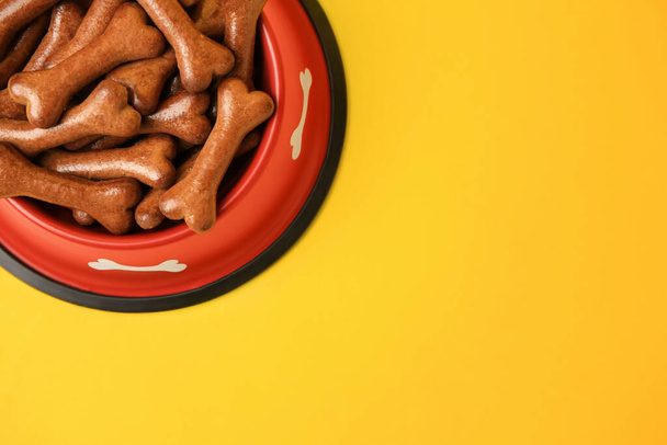 Red bowl with bone shaped dog cookies on yellow background, top view. Space for text - Photo, Image