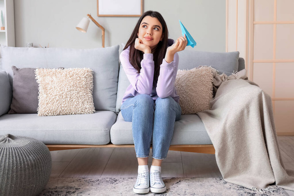 Young woman with paper plane sitting on sofa at home - Foto, Bild