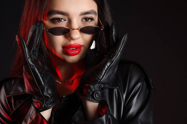 Attractive young woman in sunglasses and leather gloves on dark background, closeup - Fotoğraf, Görsel