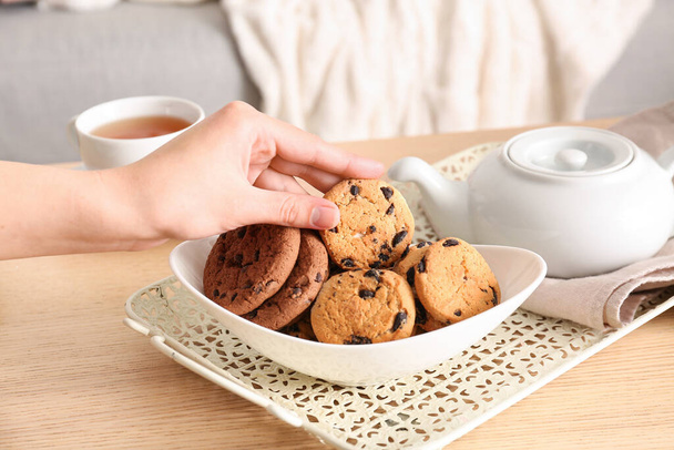Woman holding cookie and teapot with cup of tea on wooden table in living room - Foto, afbeelding