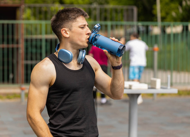 Dehydrated thirsty athletic caucasian muscular man drinking water from bottle. Tired exhausted guy on playground. Sports, health, fitness routine, workout. Runner after difficult jogging outdoors - Foto, Imagem
