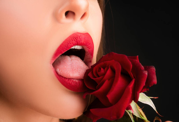 Lips with lipstick closeup. Beautiful woman lips with rose. Woman with red rose, macro, on black background - Фото, изображение