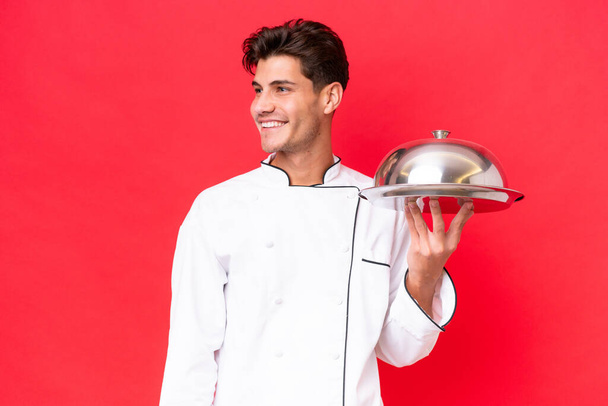 Молодий кавказький шеф-кухар тримає лоток ізольований на червоному тлі, дивлячись в бік і посміхаючись
 - Фото, зображення