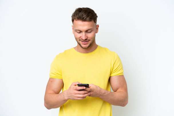 Νεαρός ξανθός καυκάσιος, απομονωμένος σε λευκό φόντο, στέλνει μήνυμα με το κινητό. - Φωτογραφία, εικόνα