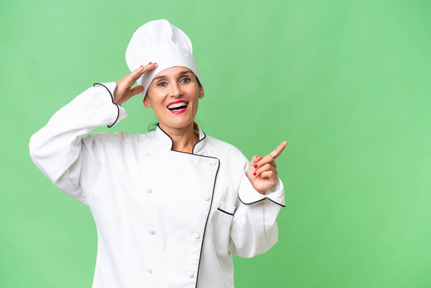Женщина-повар средних лет на изолированном фоне удивлена и указывает пальцем в сторону - Фото, изображение
