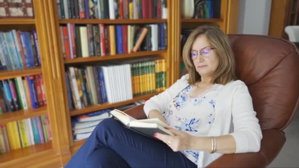 Mulher branca sênior lendo um livro sentado em uma poltrona relaxante com uma estante atrás dela - Filmagem, Vídeo