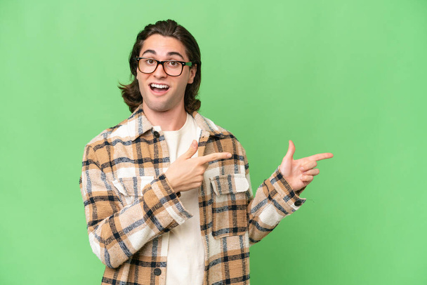 Young caucasian man isolated on green chroma background surprised and pointing side - Photo, Image