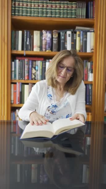 Valkoinen nainen lukee kirjaa lasipöydällä, jonka takana on hyllyllinen kirjoja - Materiaali, video