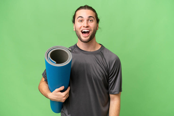 Junger Sportler besucht Yoga-Kurse, während er eine Matte mit überraschendem Gesichtsausdruck über isoliertem Hintergrund hält - Foto, Bild